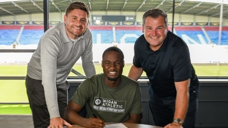 Christian Saydee, wearing a Wigan Athletic shirt, poses with two club officials after signing a three-year contract at the Brick Community Stadium ahead of the 2025–26 League One season.
