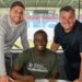 Christian Saydee, wearing a Wigan Athletic shirt, poses with two club officials after signing a three-year contract at the Brick Community Stadium ahead of the 2025–26 League One season.