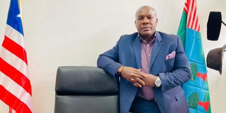 Senator Jonathon Boycharles Sogbie of River Gee County standing beside an executive chair, flanked by the Liberian national flag and River Gee County flag, after introducing a landmark sports development bill.