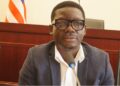 Tarnue N. Jeke, nominee for Deputy Minister for Administration at Liberia’s Ministry of Commerce, seated at a confirmation hearing before the Senate Committee, wearing a dark blazer and glasses.