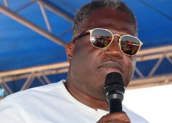 Vice President Jeremiah Koung speaks into a microphone while wearing sunglasses and a white shirt, standing under a blue canopy during a public event.