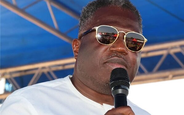 Vice President Jeremiah Koung speaks into a microphone while wearing sunglasses and a white shirt, standing under a blue canopy during a public event.