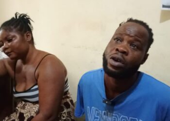 A man and a woman, identified as Zarwu Kennedy, 32, and Princess Varnah, 30, sit indoors after their arrest in Bong County, Liberia, for alleged drug trafficking. The woman appears distressed, resting her head on her hand, while the man looks upward, speaking.