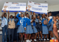 Students from Kings and Queens International School celebrate winning first place in the YMA National Press Club Exhibition, holding L$25,000 and L$40,000 championship checks on stage