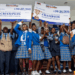 Students from Kings and Queens International School celebrate winning first place in the YMA National Press Club Exhibition, holding L$25,000 and L$40,000 championship checks on stage