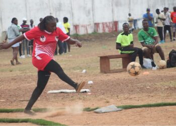 A female kickball player in red uniform kicks the ball during a dusty league match in Liberia, highlighting the sport’s resilience amid neglect