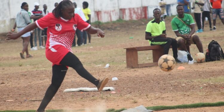 A female kickball player in red uniform kicks the ball during a dusty league match in Liberia, highlighting the sport’s resilience amid neglect