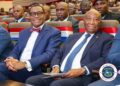 Liberian President Joseph Nyuma Boakai and African Development Bank President Dr. Akinwumi Adesina sit side by side at a high-level government event in Monrovia, surrounded by officials and dignitaries, with red, white, and blue decorations symbolizing national pride.