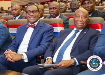 Liberian President Joseph Nyuma Boakai and African Development Bank President Dr. Akinwumi Adesina sit side by side at a high-level government event in Monrovia, surrounded by officials and dignitaries, with red, white, and blue decorations symbolizing national pride.