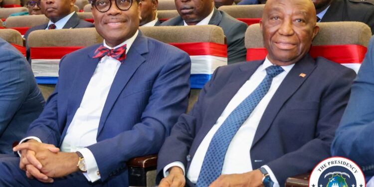 Liberian President Joseph Nyuma Boakai and African Development Bank President Dr. Akinwumi Adesina sit side by side at a high-level government event in Monrovia, surrounded by officials and dignitaries, with red, white, and blue decorations symbolizing national pride.