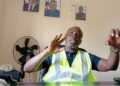 Harper Port Director Augustine Nuefville speaking in office with high-visibility vest and Liberian presidential portraits on the wall
