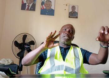 Harper Port Director Augustine Nuefville speaking in office with high-visibility vest and Liberian presidential portraits on the wall
