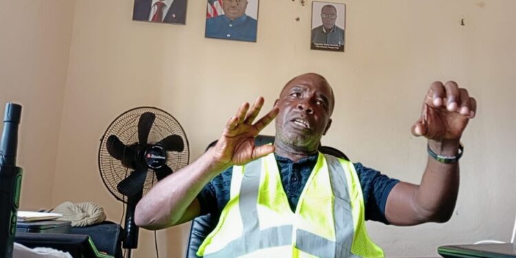 Harper Port Director Augustine Nuefville speaking in office with high-visibility vest and Liberian presidential portraits on the wall