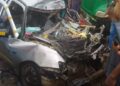 Damaged silver vehicle mangled in a fatal crash with a Bea Mountain supply truck on the Ibrahim Banbagada Highway, as bystanders gather and police investigate.