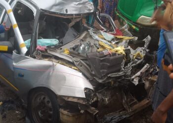 Damaged silver vehicle mangled in a fatal crash with a Bea Mountain supply truck on the Ibrahim Banbagada Highway, as bystanders gather and police investigate.
