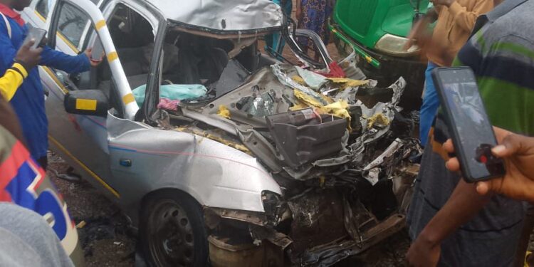 Damaged silver vehicle mangled in a fatal crash with a Bea Mountain supply truck on the Ibrahim Banbagada Highway, as bystanders gather and police investigate.