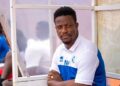 Coach Mohamed Kallon seated on the bench during a Sierra Leone national team match, wearing team gear with initials “MK.”
