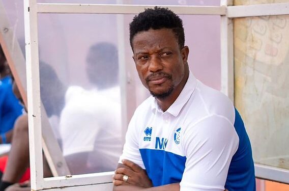 Coach Mohamed Kallon seated on the bench during a Sierra Leone national team match, wearing team gear with initials “MK.”