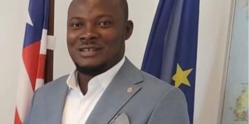 Township Commissioner Edward Horton Lahai stands near national and EU flags, highlighting governance challenges faced by local officials across Liberia.