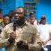Rep. James Kolleh speaks into a microphone during a public gathering at Radio Gbartala's 13th anniversary celebration, surrounded by community members and supporters in Bong County, Liberia.