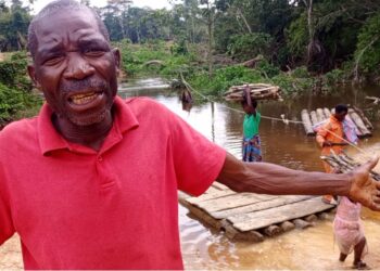 Frustrated elder in Loguatuo, Nimba County, pleads for bridge construction over the deadly Louhn River as residents cross on a makeshift raft in the background