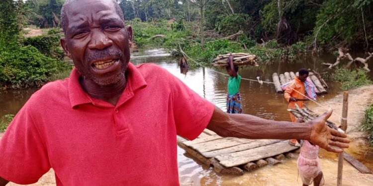 Frustrated elder in Loguatuo, Nimba County, pleads for bridge construction over the deadly Louhn River as residents cross on a makeshift raft in the background