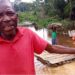 Frustrated elder in Loguatuo, Nimba County, pleads for bridge construction over the deadly Louhn River as residents cross on a makeshift raft in the background