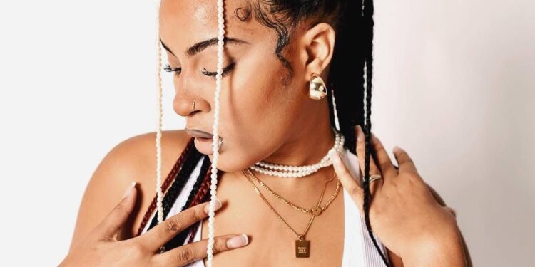 Liberian socialite Michelle Doe poses in a white tank top with intricately braided hair adorned with beads and pearls, symbolizing her strength and individuality as highlighted in her public journey of resilience and reinvention