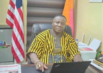 Rep. Prince Koinah seated at his office desk, wearing traditional Liberian attire, amid growing backlash from Kpaai Town residents over stalled campaign projects and poor road conditions.