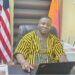 Rep. Prince Koinah seated at his office desk, wearing traditional Liberian attire, amid growing backlash from Kpaai Town residents over stalled campaign projects and poor road conditions.