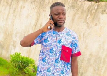 Julius Sevelee, the teacher suspended in Bong County, stands on his phone near a school compound.