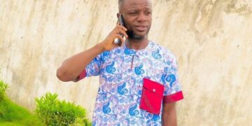 Julius Sevelee, the teacher suspended in Bong County, stands on his phone near a school compound.