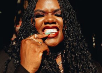 Liberian artist Ceekaynayway smiles boldly in a close-up portrait with braided hair, gold rings, and red lipstick, embodying confidence and authenticity