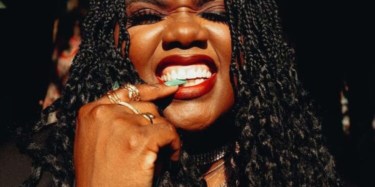 Liberian artist Ceekaynayway smiles boldly in a close-up portrait with braided hair, gold rings, and red lipstick, embodying confidence and authenticity