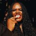 Liberian artist Ceekaynayway smiles boldly in a close-up portrait with braided hair, gold rings, and red lipstick, embodying confidence and authenticity