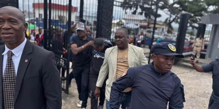 Fobay Emery Johnson, alleged Ecobank robbery suspect, escorted by police officers outside the Monrovia City Court gate after being charged with armed robbery and attempted murder.