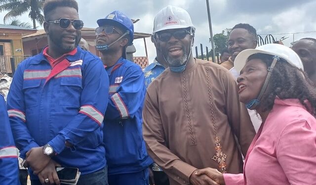 Representative Jeremiah Sokan Sr. in a brown kaftan greets a female engineer during a site visit in Grand Gedeh County, surrounded by local workers and project staff.