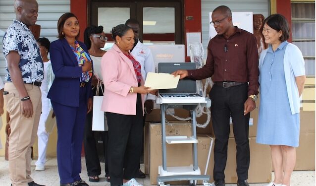 Dr. Teyah S. Moore hands over documents for new ultrasound machines to JFK CEO Dr. Linda Birch, with Ministry of Health and hospital staff present.