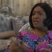 Former Liberian Vice President Jewel Howard-Taylor gestures expressively during an interview, seated indoors with a Liberian flag in the background and a decorative sofa behind her.
