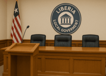 A government meeting room in Liberia featuring a wooden podium with a microphone, three empty black leather chairs behind a long wooden table, the Liberian flag in the corner, and a circular emblem on the wall reading "Liberia Local Government."