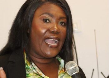 Rochell Woodson speaks into a microphone during a public address, wearing a floral blouse and black blazer.