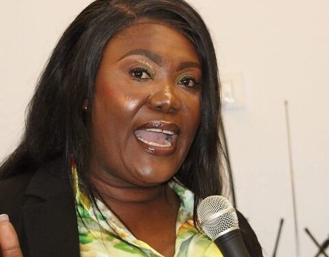Rochell Woodson speaks into a microphone during a public address, wearing a floral blouse and black blazer.
