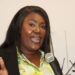 Rochell Woodson speaks into a microphone during a public address, wearing a floral blouse and black blazer.