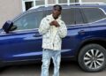 SIO standing in front of a blue SUV, wearing a patterned jacket and jeans — representing the Liberian artist’s public image amid recent controversies.