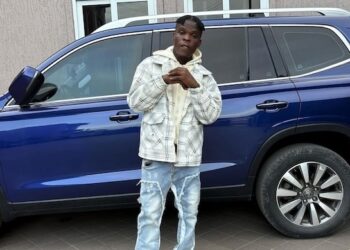SIO standing in front of a blue SUV, wearing a patterned jacket and jeans — representing the Liberian artist’s public image amid recent controversies.