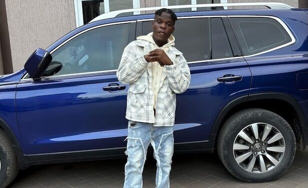 SIO standing in front of a blue SUV, wearing a patterned jacket and jeans — representing the Liberian artist’s public image amid recent controversies.