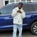 SIO standing in front of a blue SUV, wearing a patterned jacket and jeans — representing the Liberian artist’s public image amid recent controversies.