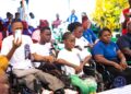 A group of persons with disabilities, including children and adults, attentively participate in an outdoor event hosted by the National Commission on Disabilities in Liberia. Some attendees are seated in wheelchairs, wearing white or blue t-shirts branded with the NCD logo, while others are dressed casually. A man in the front row records with his smartphone as a photographer captures the moment in the background. The gathering highlights community engagement and inclusion.