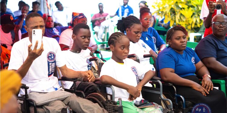 A group of persons with disabilities, including children and adults, attentively participate in an outdoor event hosted by the National Commission on Disabilities in Liberia. Some attendees are seated in wheelchairs, wearing white or blue t-shirts branded with the NCD logo, while others are dressed casually. A man in the front row records with his smartphone as a photographer captures the moment in the background. The gathering highlights community engagement and inclusion.