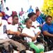 A group of persons with disabilities, including children and adults, attentively participate in an outdoor event hosted by the National Commission on Disabilities in Liberia. Some attendees are seated in wheelchairs, wearing white or blue t-shirts branded with the NCD logo, while others are dressed casually. A man in the front row records with his smartphone as a photographer captures the moment in the background. The gathering highlights community engagement and inclusion.